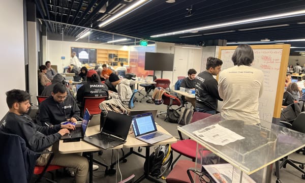 Our team at MIT iQuHACK working on laptops at tables, with a whiteboard covered in quantum challenge notes and other hackers in the background.