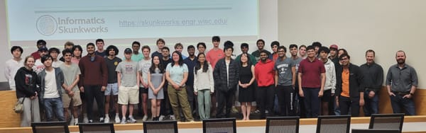 Informatics Skunkworks research group all hands meeting with members posing in front of a projector screen displaying the group logo and website.