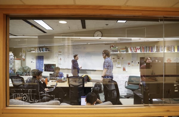 View through the UPL window of Andrew and Ben standing at the whiteboard while Nico talks to Phoenix on the far right, with other members working at laptops around the table.