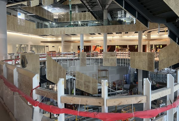 Interior of Morgridge Hall under construction with multiple open floors, exposed structural beams, perforated metal panels, and workers in safety gear.