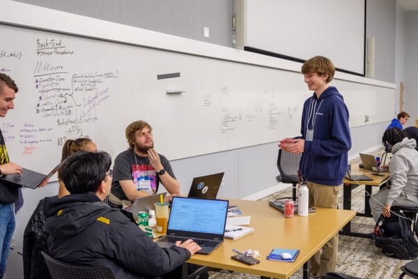Me mentoring a team at MadHacks, with the team clustered around a couple computers, and a marked up whiteboard behind them.