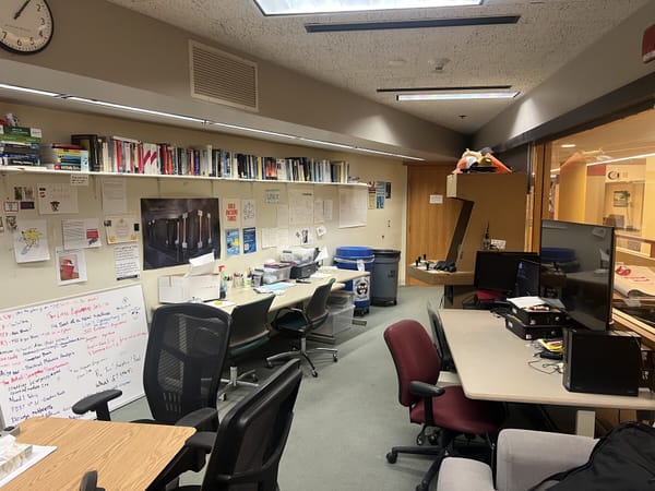 Modern photo of the UPL, featuring workstations, whiteboards covered in notes, bookshelves, and a clock on the wall.